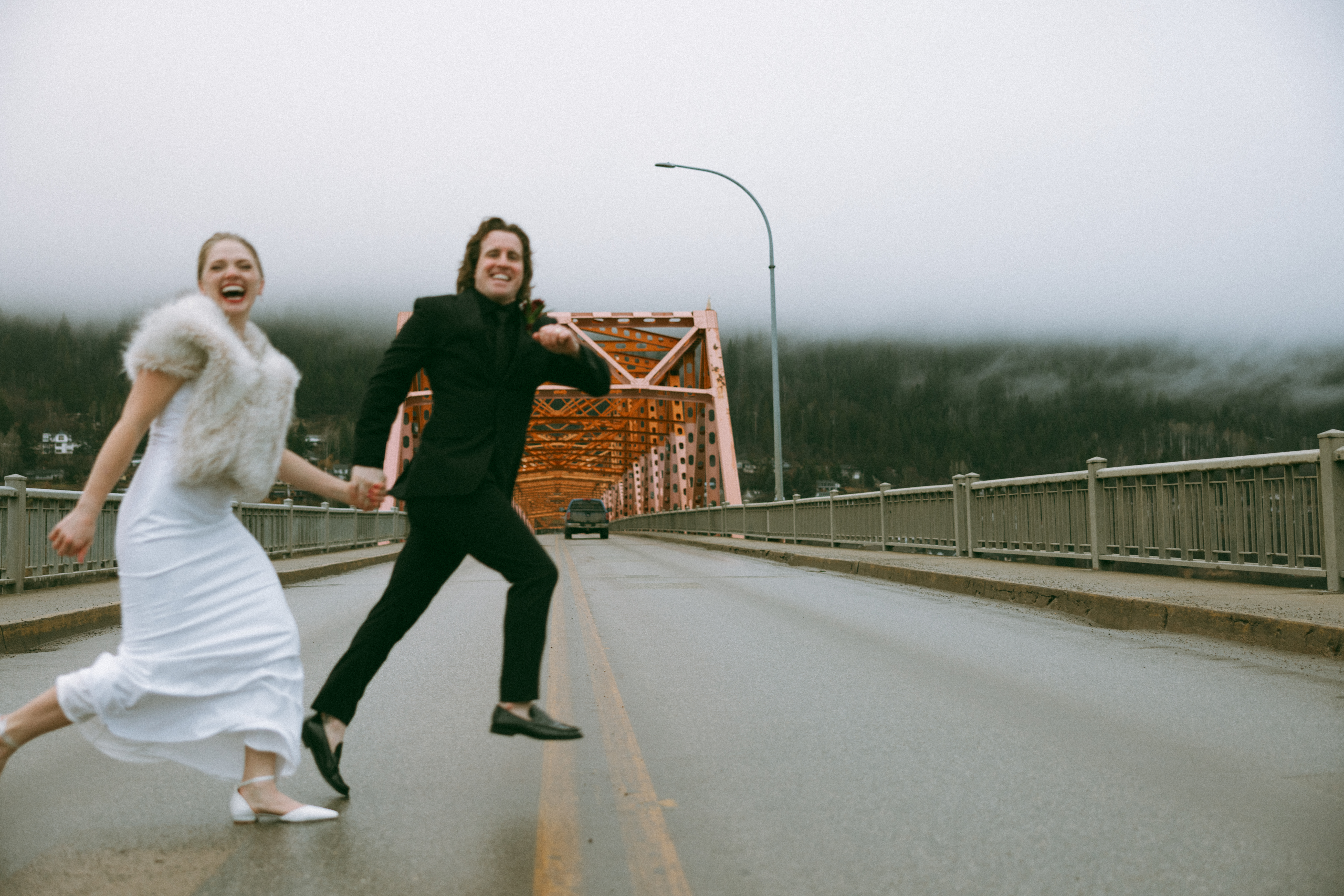 A bride and groom hold hands while running playfully across a road, with a big orange bridge in the background. West Kootenay Wedding Photographer Nelson BC Elopement